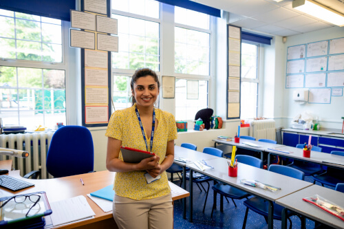 Teacher with clip board leaning against a desk in a school